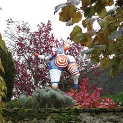 Nana rouge et bleue en situation au dessus d'un mur entre un cotinus rouge et des santolines grises mettant en valeur les couleurs vives de la sculpture